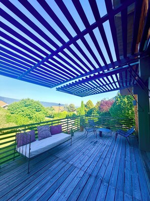 Eine Holzterrasse mit einer Pergola, Sitzgelegenheiten im Freien und Blick auf Grünflächen und Hügel.