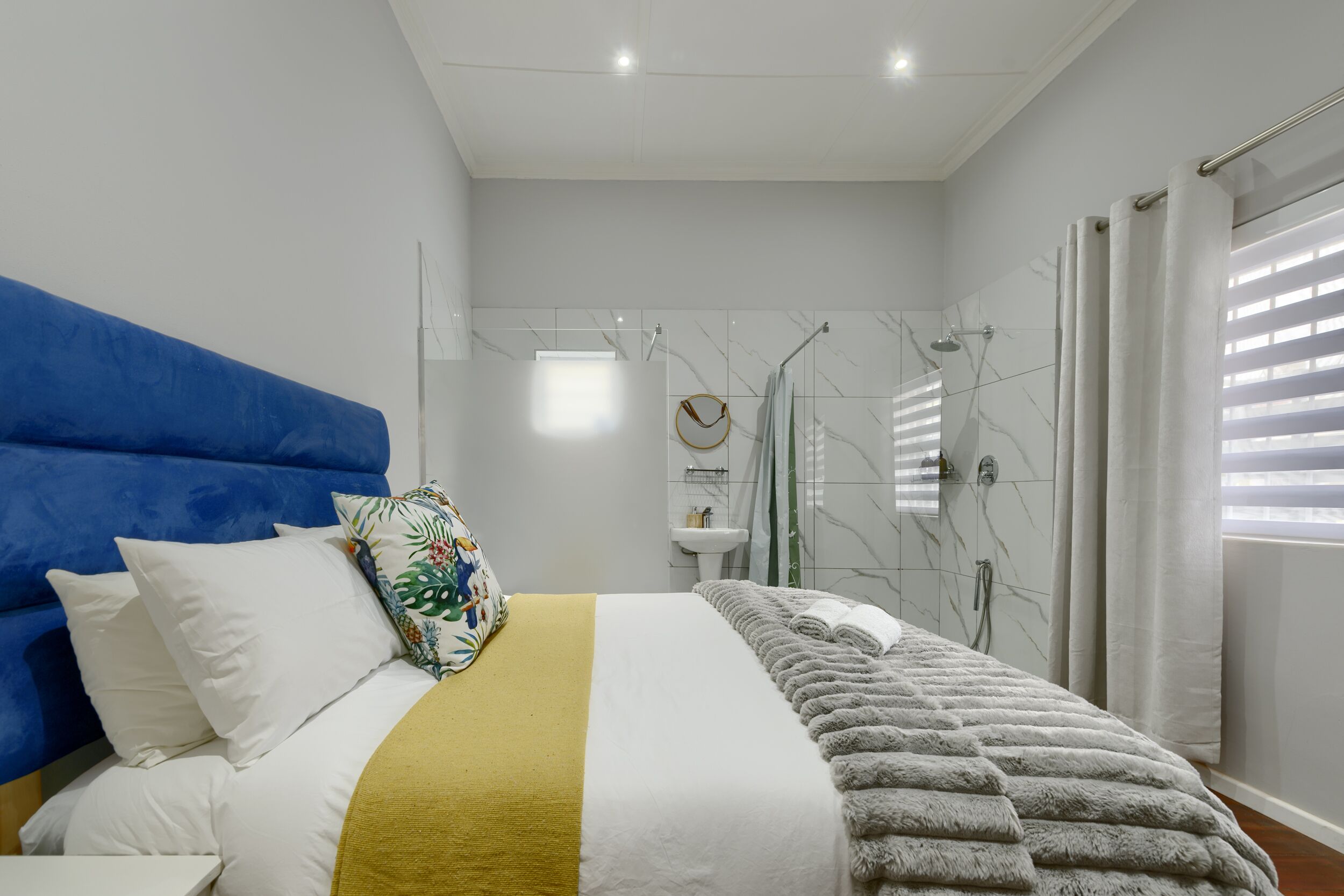 A modern bedroom with a blue headboard, a bed with white and yellow bedding, a bathroom with a shower, and a window with blinds.