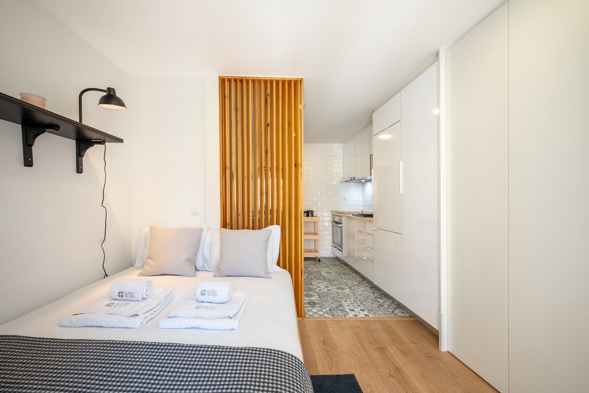 A modern hotel room with a bed, a desk, a lamp, and a view into the kitchen.