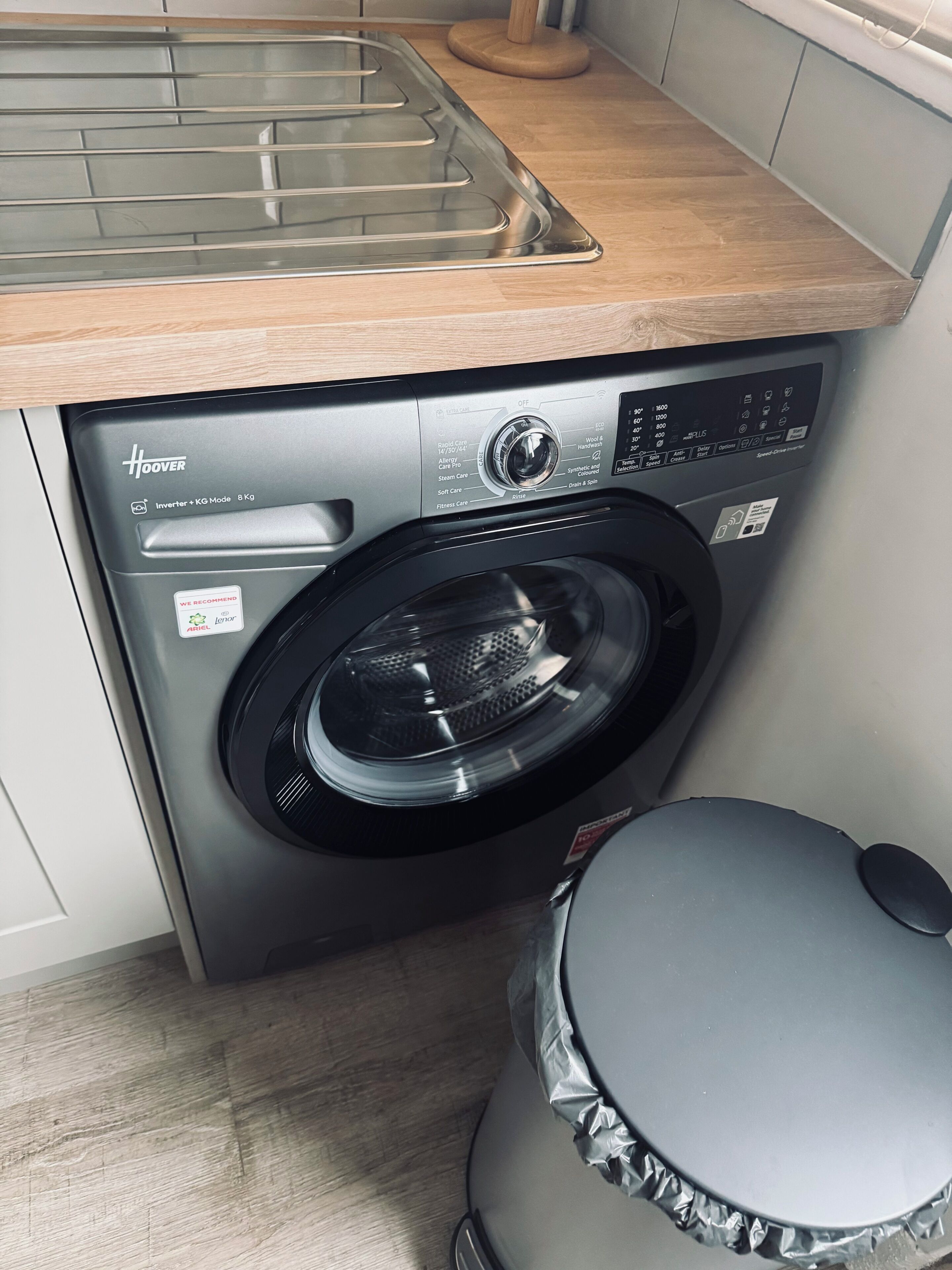 Una lavadora compacta y moderna con panel de control, ubicada en un pequeño cuarto de lavado con una encimera de madera.