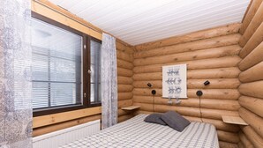 A log cabin bedroom with a bed, a window with blinds, a radiator, and a wall-mounted light fixture. - Onnela 1 Chalet | Sauna & Ski Trails | Ruka (Kuusamo)