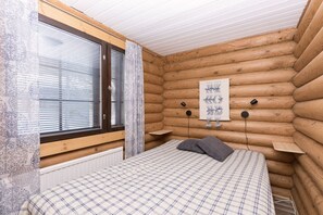 Une chambre dans un chalet en rondins, avec un lit, une fenêtre avec des stores, un radiateur et un luminaire fixé au mur.
