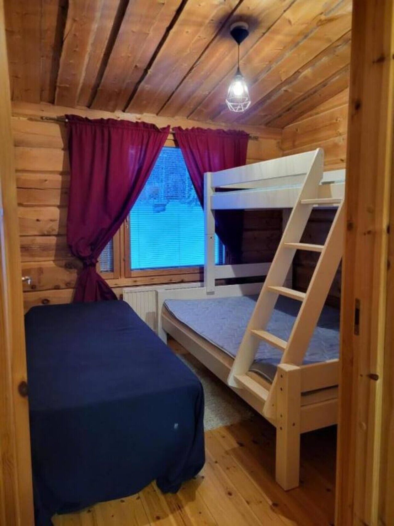 A wooden cabin room with bunk beds, a ladder, a window with curtains, and a ceiling light fixture.