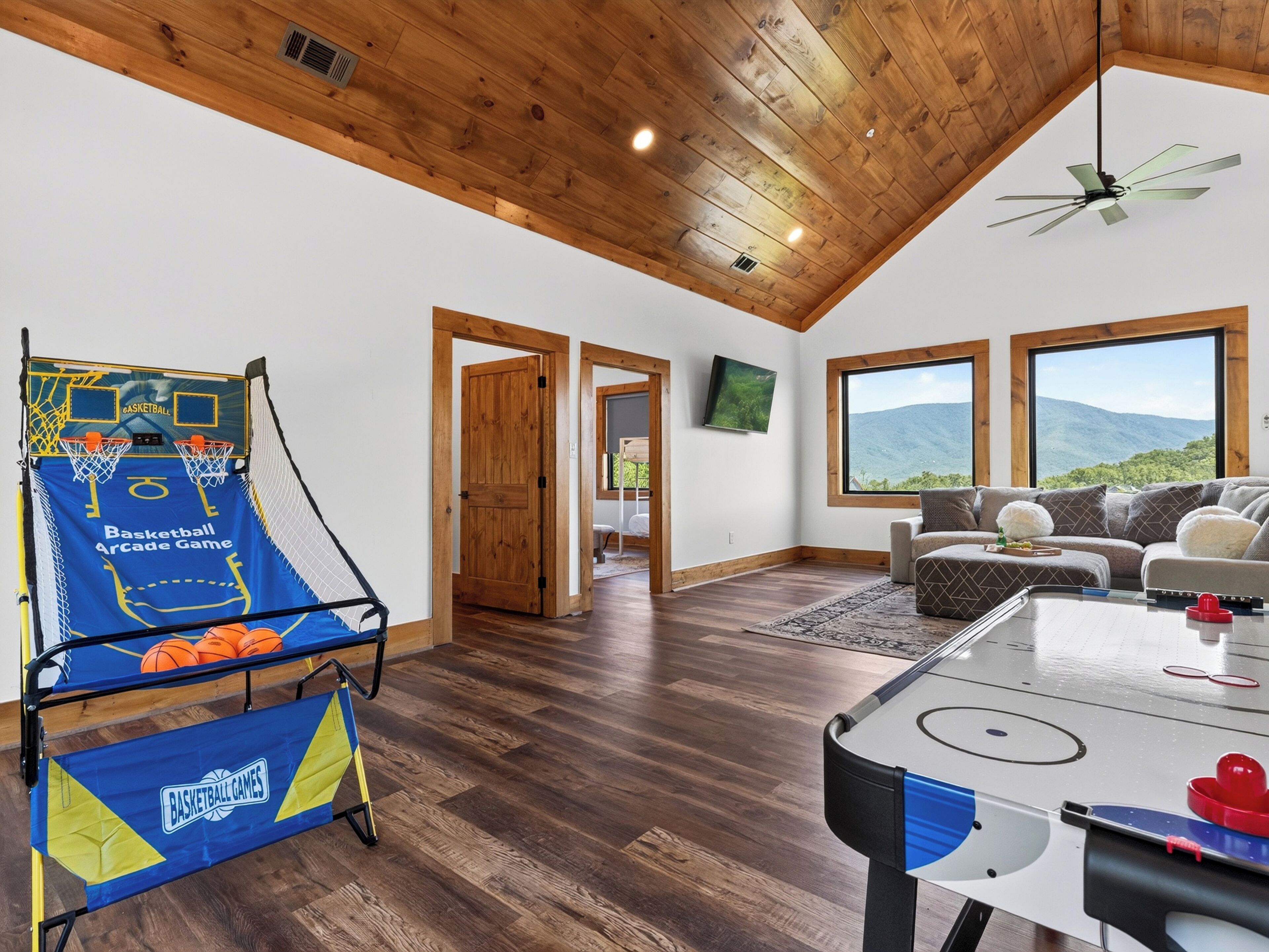 A spacious room with a basketball arcade game, a ceiling fan, a large window with a mountain view, and a cozy seating area.