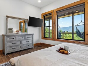 A bedroom with a large bed, a dresser, a flat-screen TV, and a window with a view of greenery and mountains. - Sleeps 40 + 2 Indoor Pools + Game Rooms | The Spurling Collection (Sevierville)
