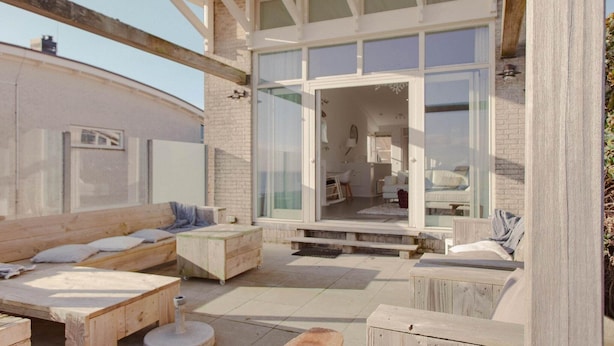 A modern outdoor living space with a wooden deck, seating area, and a glass door leading to an interior room. - Villa with Panoramic Water Views (Makkum)