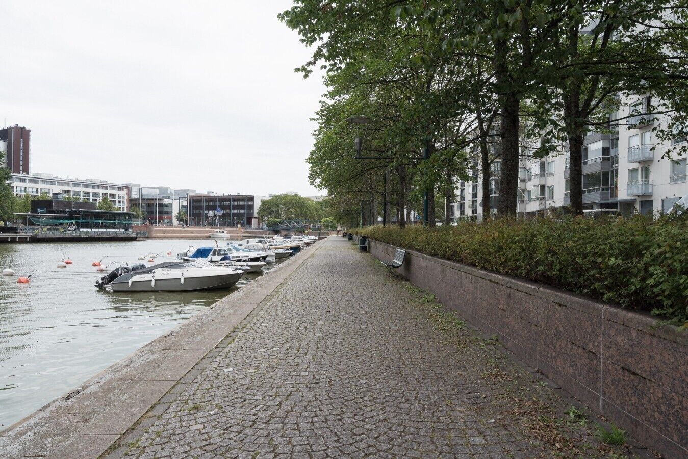A waterfront walkway with boats docked on the left, lined with trees and a stone wall on the right.