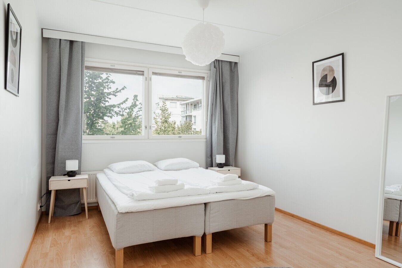 A modern bedroom with a large bed, two bedside tables, a window with curtains, and a hanging light fixture.