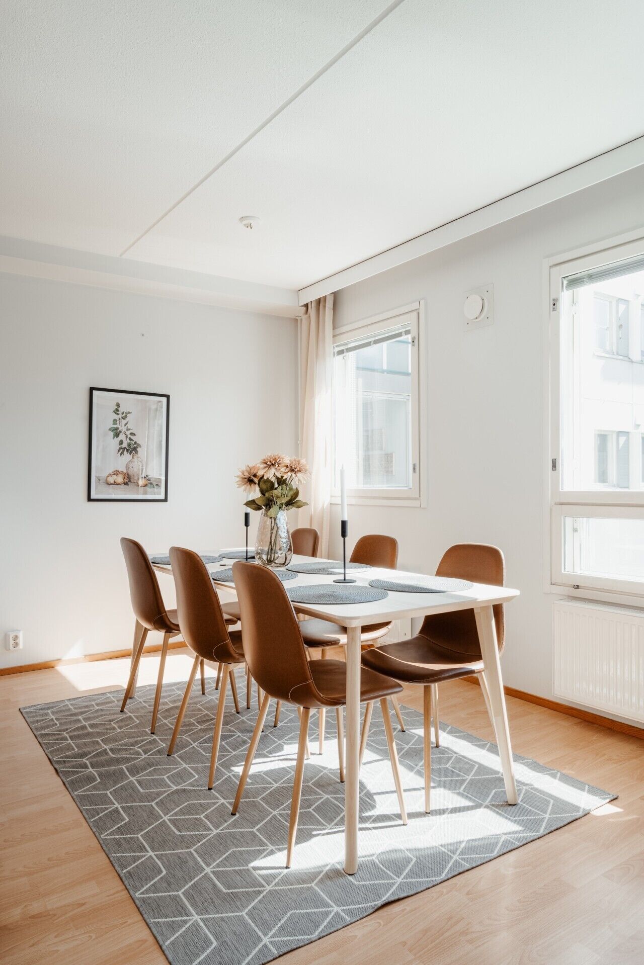 A modern dining area with a round table, six chairs, a vase with flowers, and a framed picture on the wall.