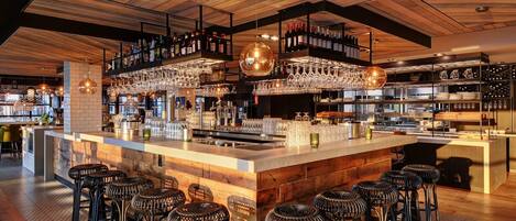 Zona de bar con techo de madera, taburetes y estantes llenos de botellas y copas.