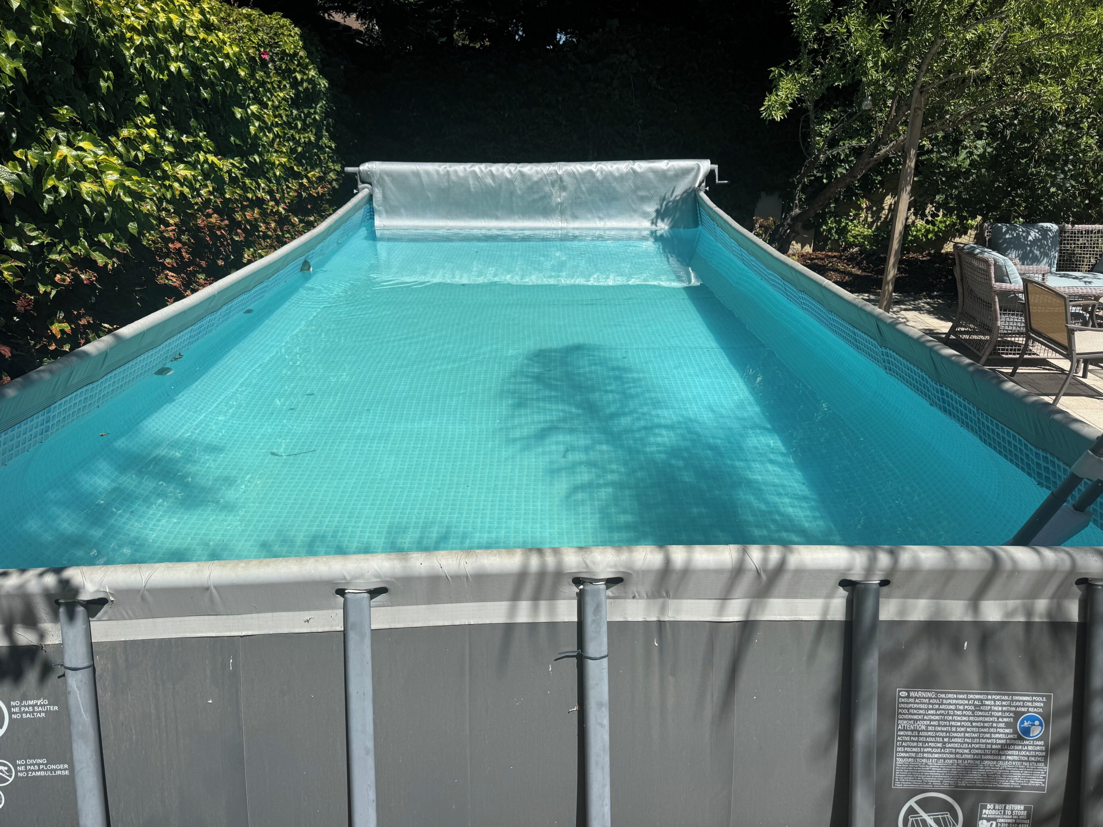 Ein Schwimmbecken mit überdachtem Bereich, umgeben von Bäumen und Gartenmöbeln.