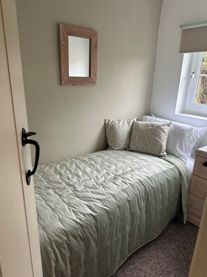 Ein Einzelbett mit gesteppter Decke, ein Nachttisch, eine Kommode, ein Fenster mit Jalousie und ein Spiegel an der Wand.