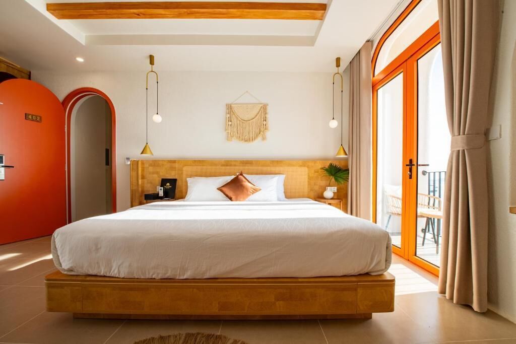 A modern hotel room with a large bed, a wooden headboard, a wall-mounted macrame decoration, and a sliding glass door to the balcony.