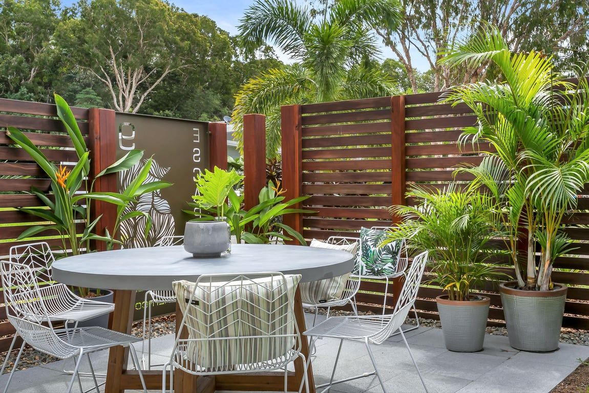 An outdoor patio with a round table and white metal chairs, surrounded by potted plants and a wooden fence.
