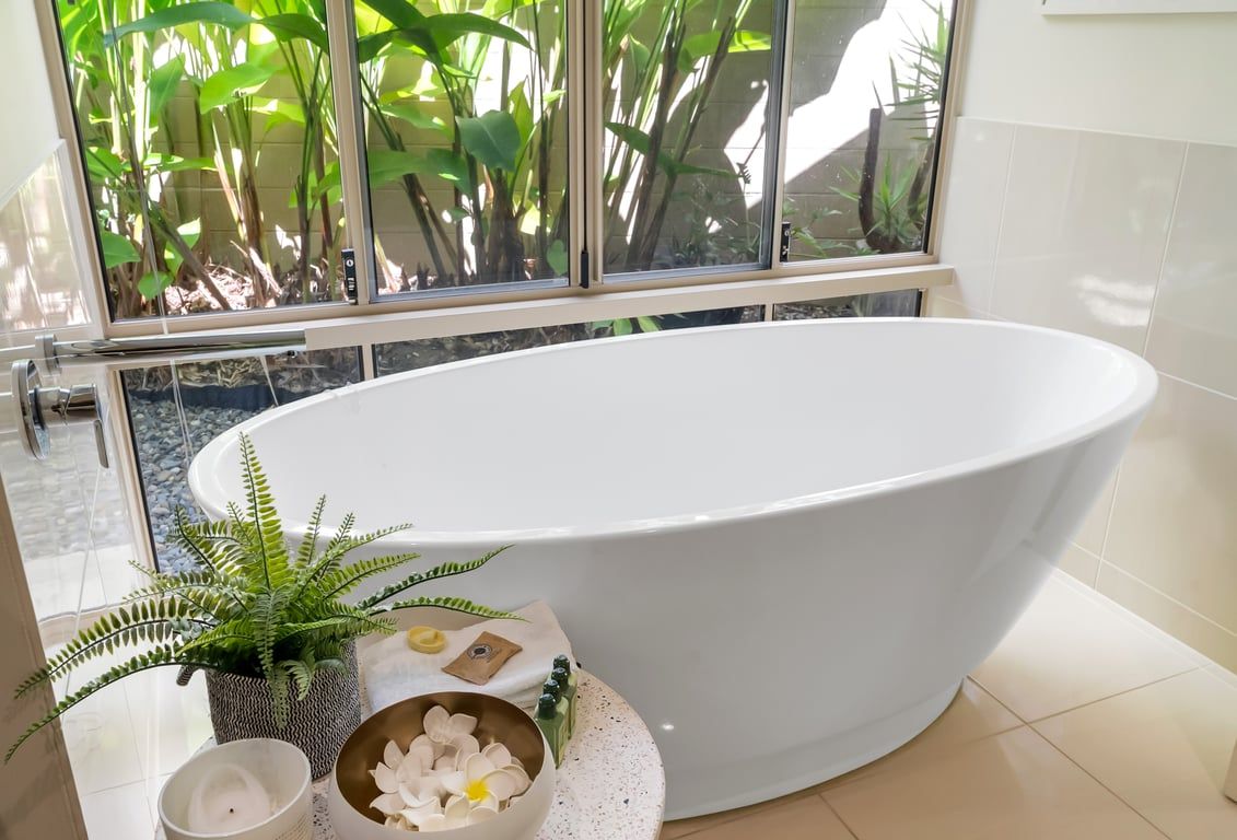 A modern bathroom with a freestanding bathtub, a potted fern, a candle, and a bowl of white flowers.