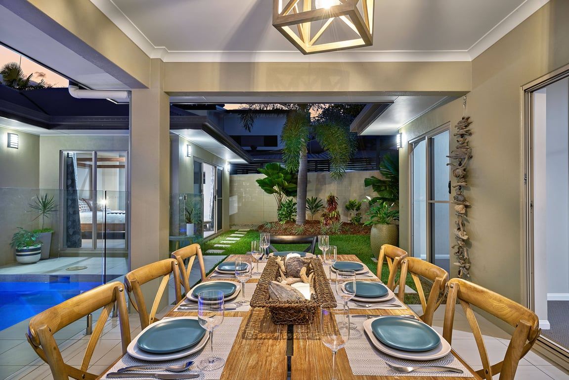 A dining area with a wooden table set for six, surrounded by wooden chairs, with a view of a swimming pool and greenery in the background.
