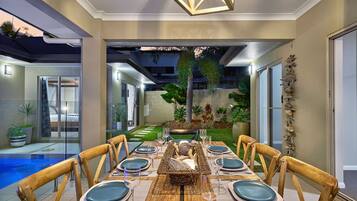 A dining area with a wooden table set for six, surrounded by wooden chairs, with a view of a swimming pool and greenery in the background.