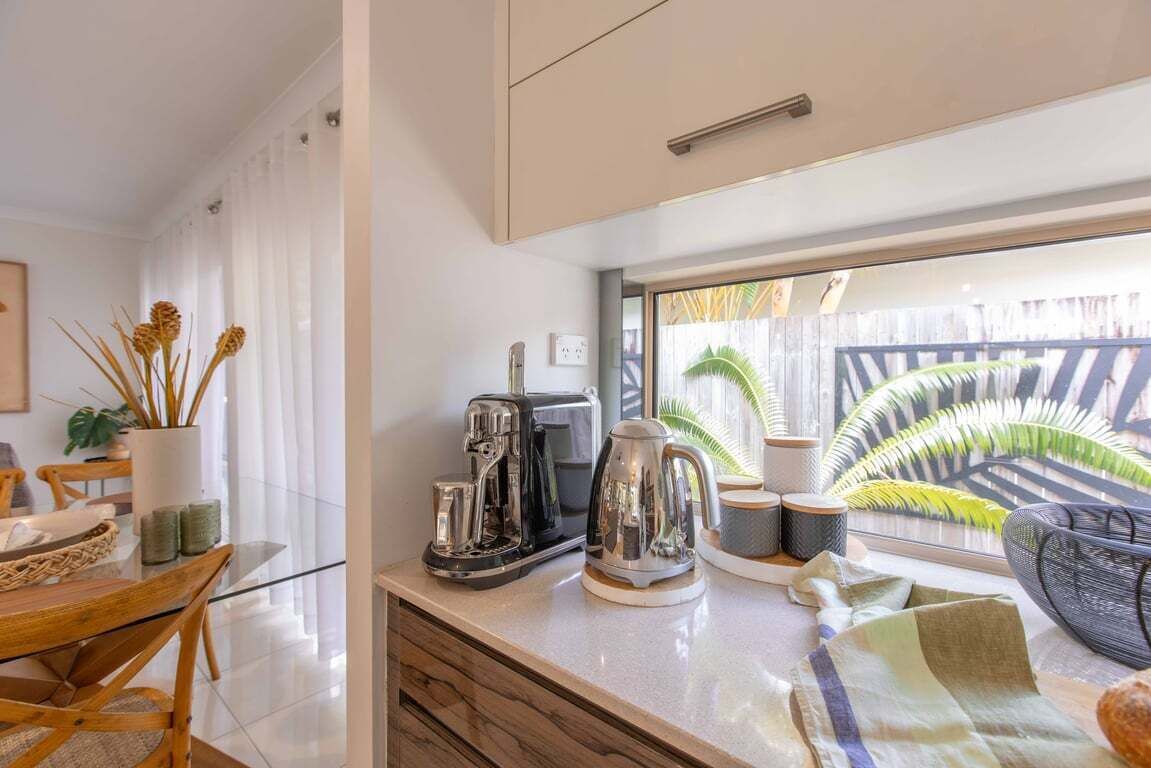 Une cuisine moderne avec une table en verre, une machine à café et une vue sur un jardin, visible à travers une fenêtre.