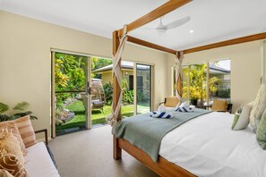 Une chambre à coucher avec un lit à baldaquin, un ventilateur de plafond et une vue sur un jardin tropical, visible à travers des portes vitrées.