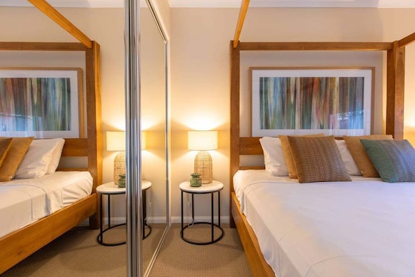 A hotel room with two beds, each with a wooden headboard, a round side table, a floor lamp, and a mirror.