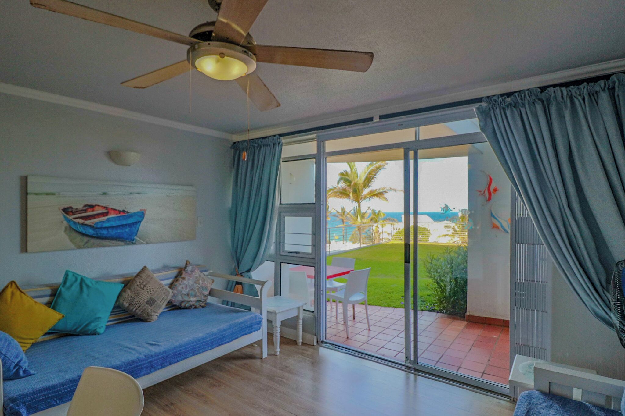 A room with a bed, a ceiling fan, a painting, and a view of a beach.