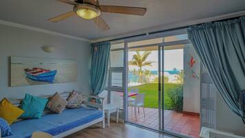 A room with a bed, a ceiling fan, a painting, and a view of a beach.