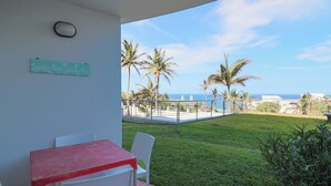 A balcony with a table and chairs, a sign reading 'Sandy Feet Welcome Here', and a view of the ocean with palm trees.