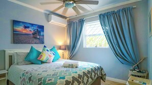 A bedroom with a bed, pillows, a nightstand, a lamp, a ceiling fan, a window with curtains, and a painting on the wall.
