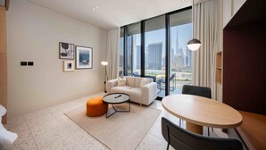 Ein modernes Hotelzimmer mit großem Fenster und Blick auf die Stadt, einer Sitzecke, einem kleinen runden Tisch sowie einem Esstisch mit Stühlen.
