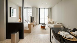 A modern hotel room with a bed, a seating area with a sofa and a coffee table, a dining table with plates and glasses, and a large window with curtains. - Furnished Business Bay Studio w/ Balcony (Dubai)