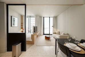 A modern hotel room with a bed, a seating area with a sofa and a coffee table, a dining table with plates and glasses, and a large window with curtains. - Furnished Business Bay Studio w/ Balcony (Dubai)