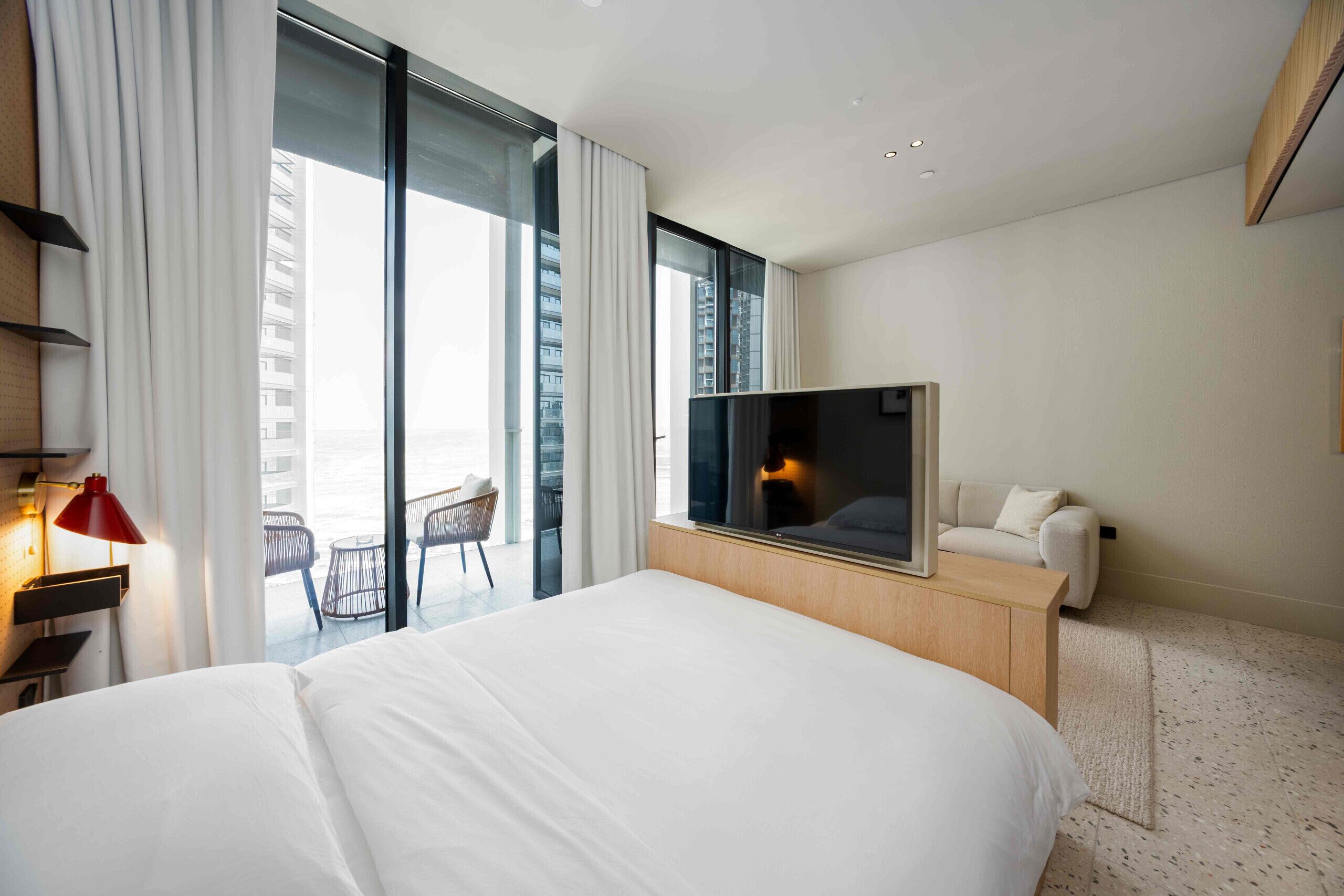A hotel room with a bed, a TV, a desk with a lamp, a chair, and a view of the city through large windows.