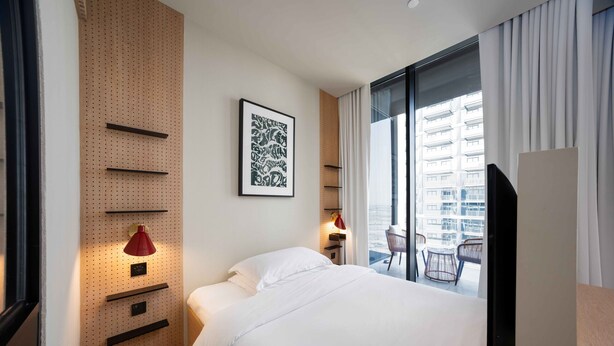A hotel room with a bed, a wall-mounted TV, a red lamp, and a window with a view of a building. - Furnished Business Bay Studio w/ Balcony (Dubai)