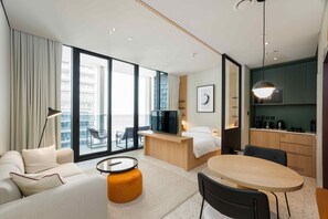 Ein modernes Hotelzimmer mit einer Couch, einem runden Tisch, einem Fernseher, einem Bett, einem Balkon mit Blick auf die Stadt und einer Küchenzeile.