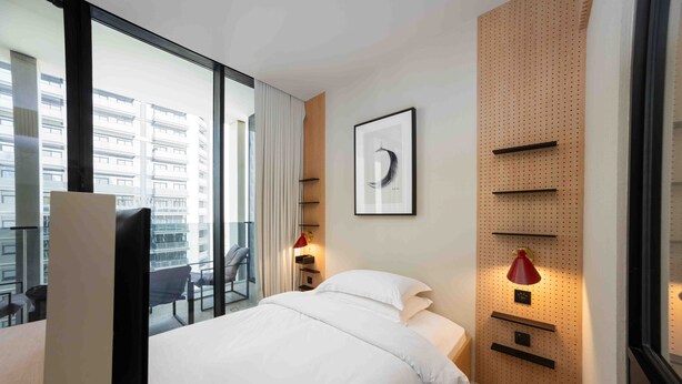 Quarto de hotel com uma cama, uma escrivaninha, uma cadeira, uma janela com vista para prédios e uma prateleira com luminária.