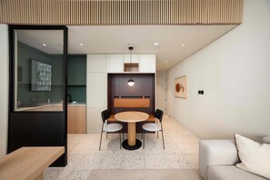 Ein modernes Interieur mit Essbereich, der einen runden Tisch und Stühle umfasst, einer Küche mit Glaskonstruktion sowie einem Wohnbereich mit Sofa und einem Wandbild.
