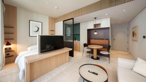Quarto de hotel moderno com cama, TV, escrivaninha, espaço de jantar e sofá.