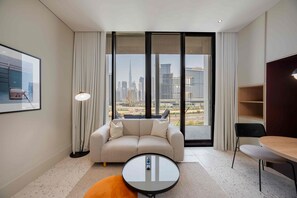 Ein modernes Hotelzimmer mit einer Couch, einem kleinen runden Tisch, einem Sessel und Blick auf eine Stadtlandschaft durch große Fenster.