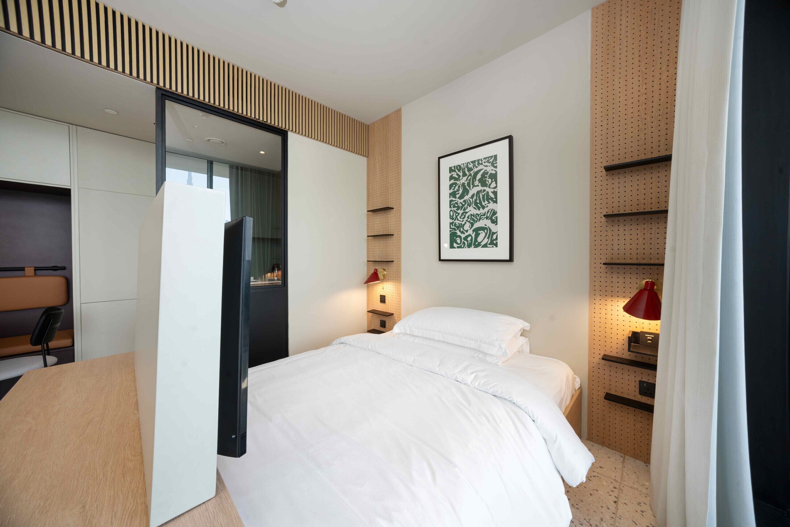 Una habitación de hotel moderna con cama, escritorio, silla, armario y estante en la pared.