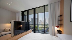 Quarto de hotel moderno com uma cama grande, televisão de tela plana, área de estar com sofá e poltronas, e varanda com vista para a cidade.