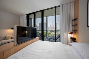 Ein modernes Hotelzimmer mit einem großen Bett, einem Flachbildfernseher, einer Sitzecke mit Sofa und Sesseln sowie einem Balkon mit Blick auf die Stadt.