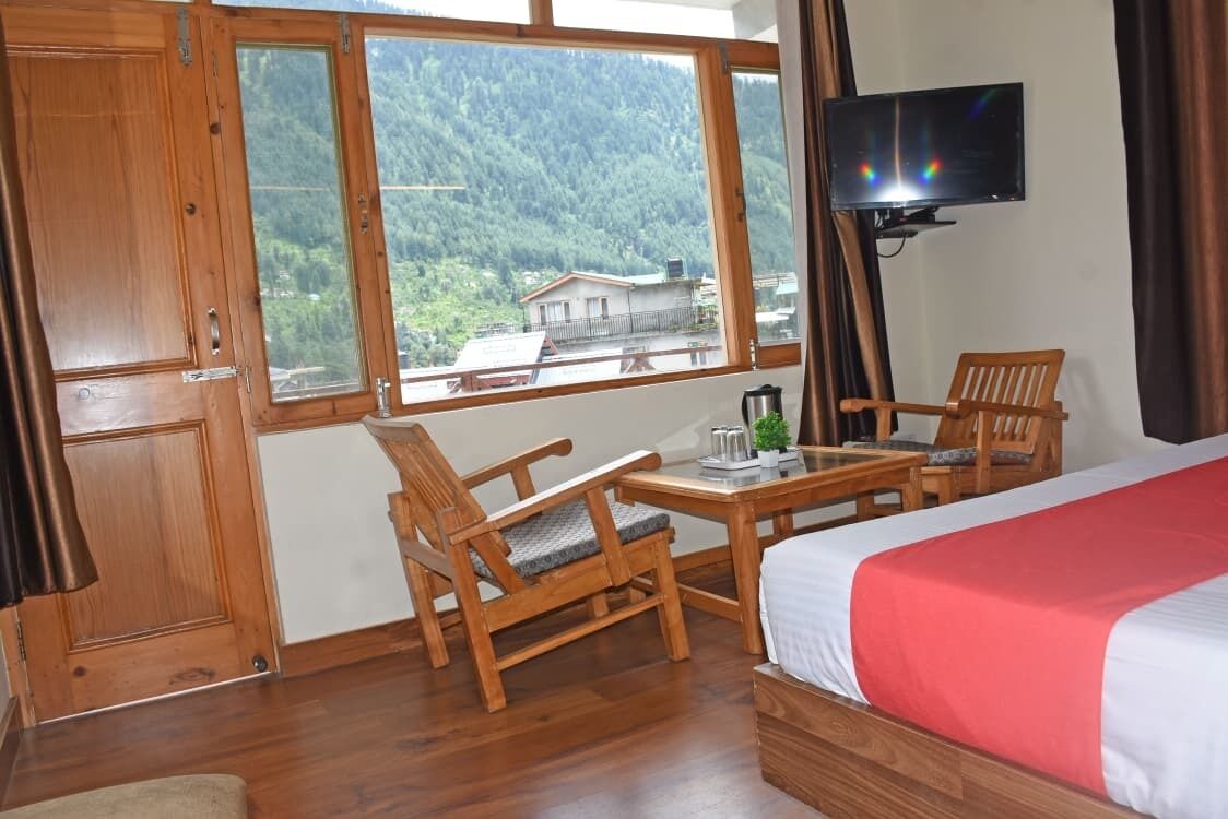 A hotel room with a bed, a desk, two chairs, a television, and a view of a mountainous landscape.