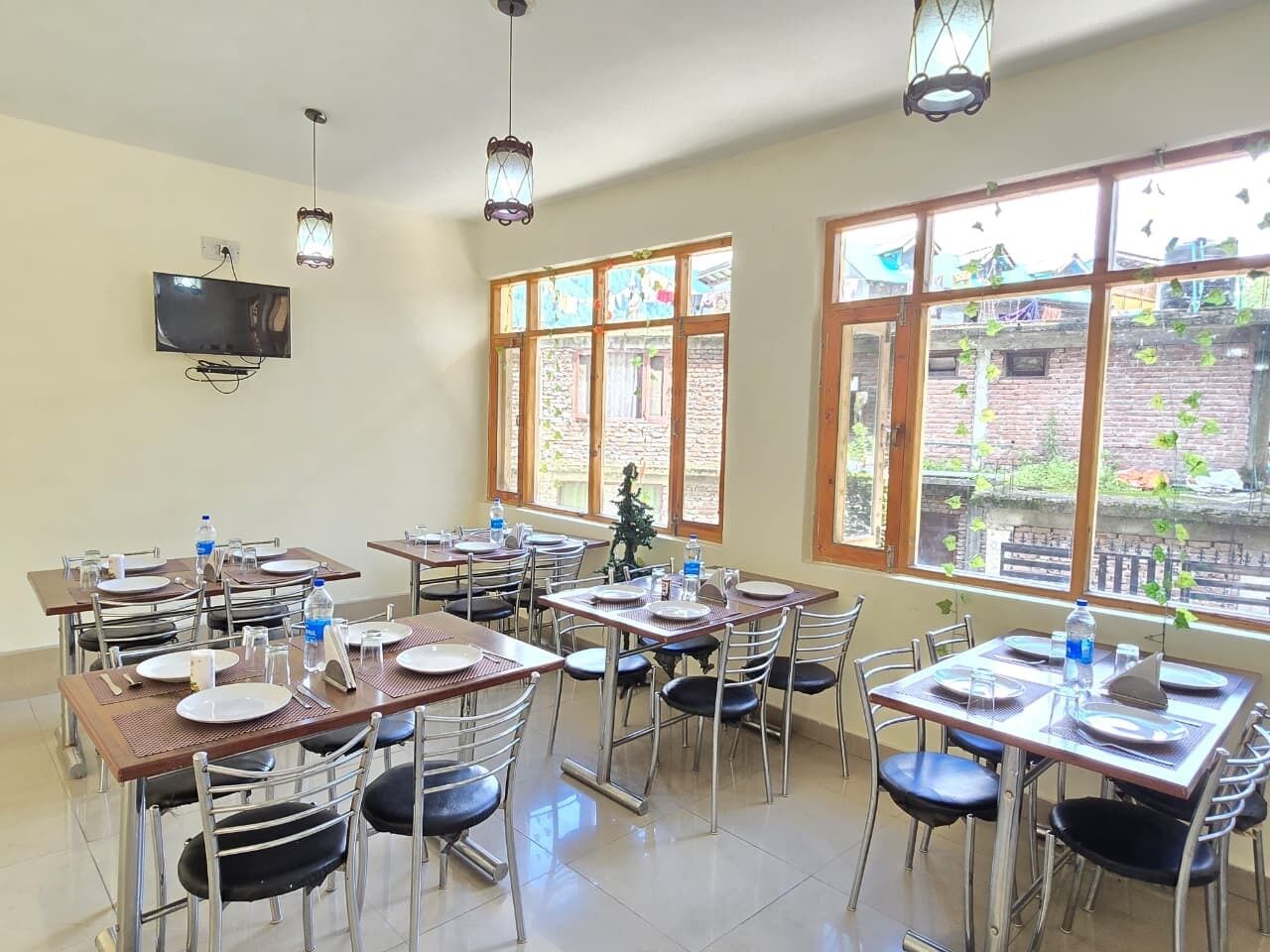 Un comedor con mesas dispuestas para comidas, un televisor montado en la pared y grandes ventanales con vistas al exterior.