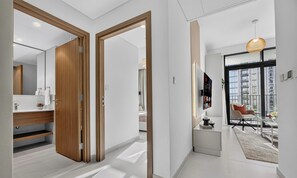 A modern interior with a wooden door leading to a bathroom, a hallway with a view of a cityscape, and a seating area with a table and chairs. - Copper & Clay 1BR - Creek Harbour (Dubai)