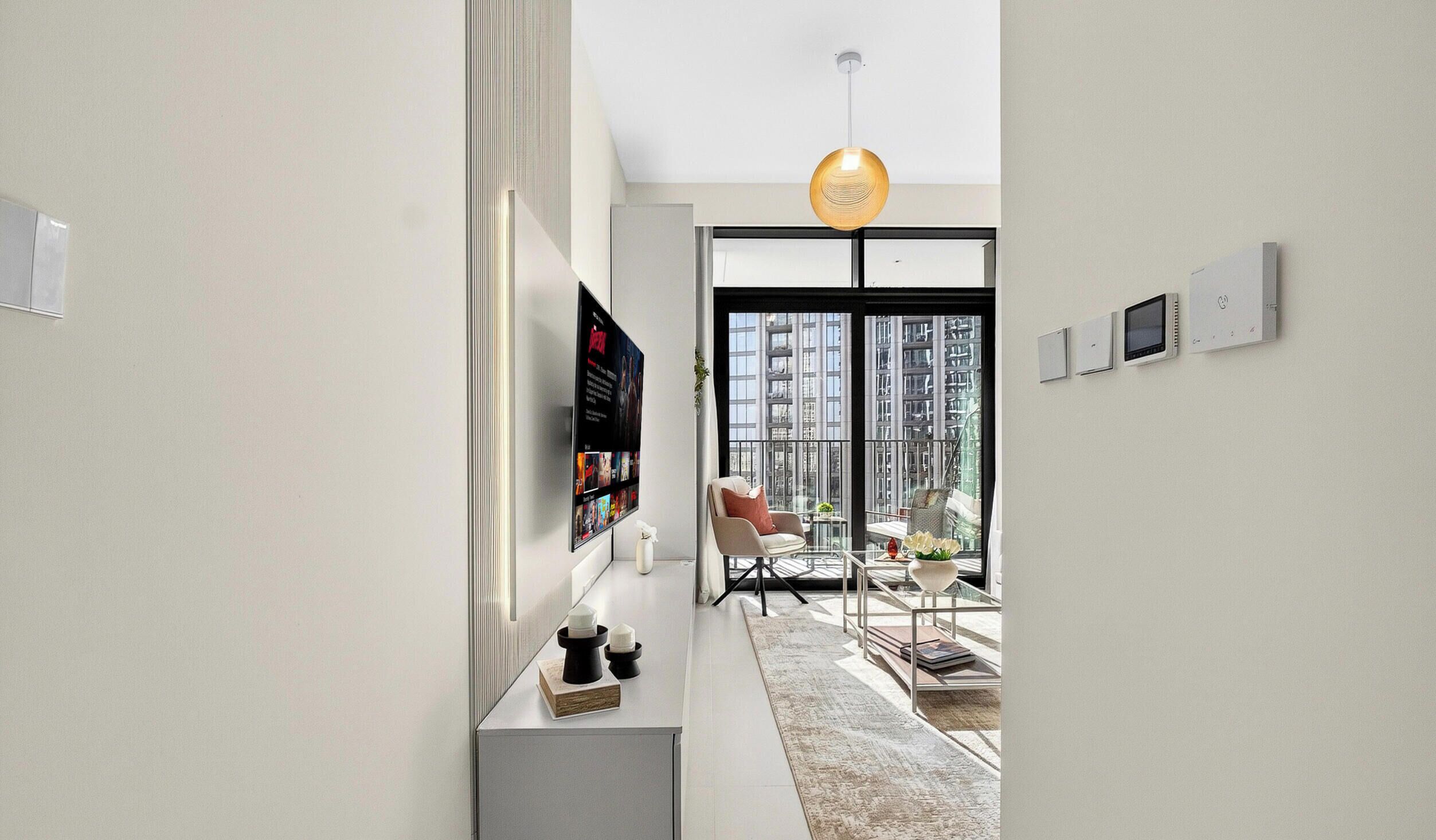 Un intérieur moderne avec un téléviseur à écran plat fixé au mur, une table basse en verre et une vue sur le paysage urbain à travers de grandes fenêtres.