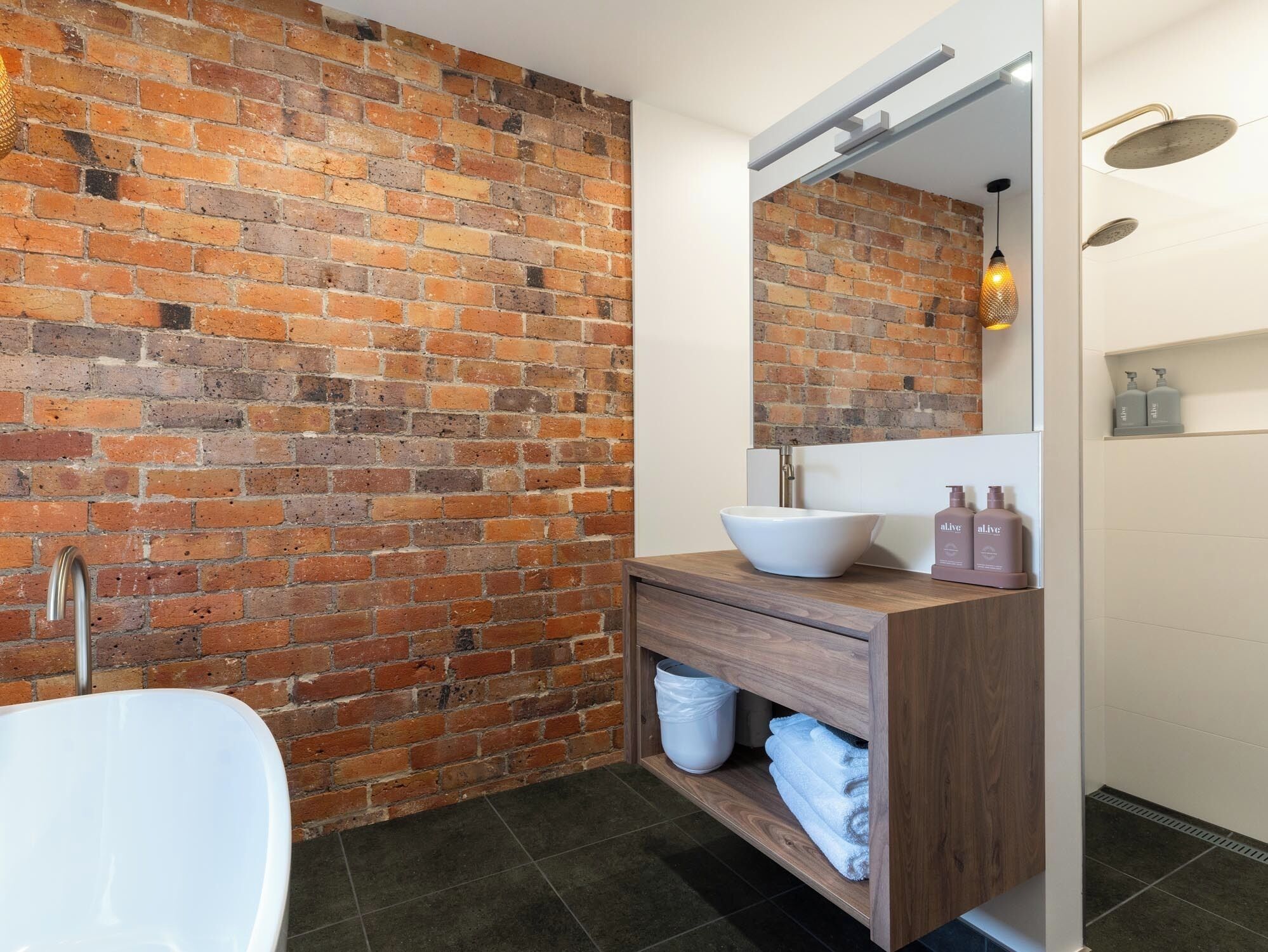 A bathroom with a white bathtub, a wooden vanity with a large mirror, and a brick wall.