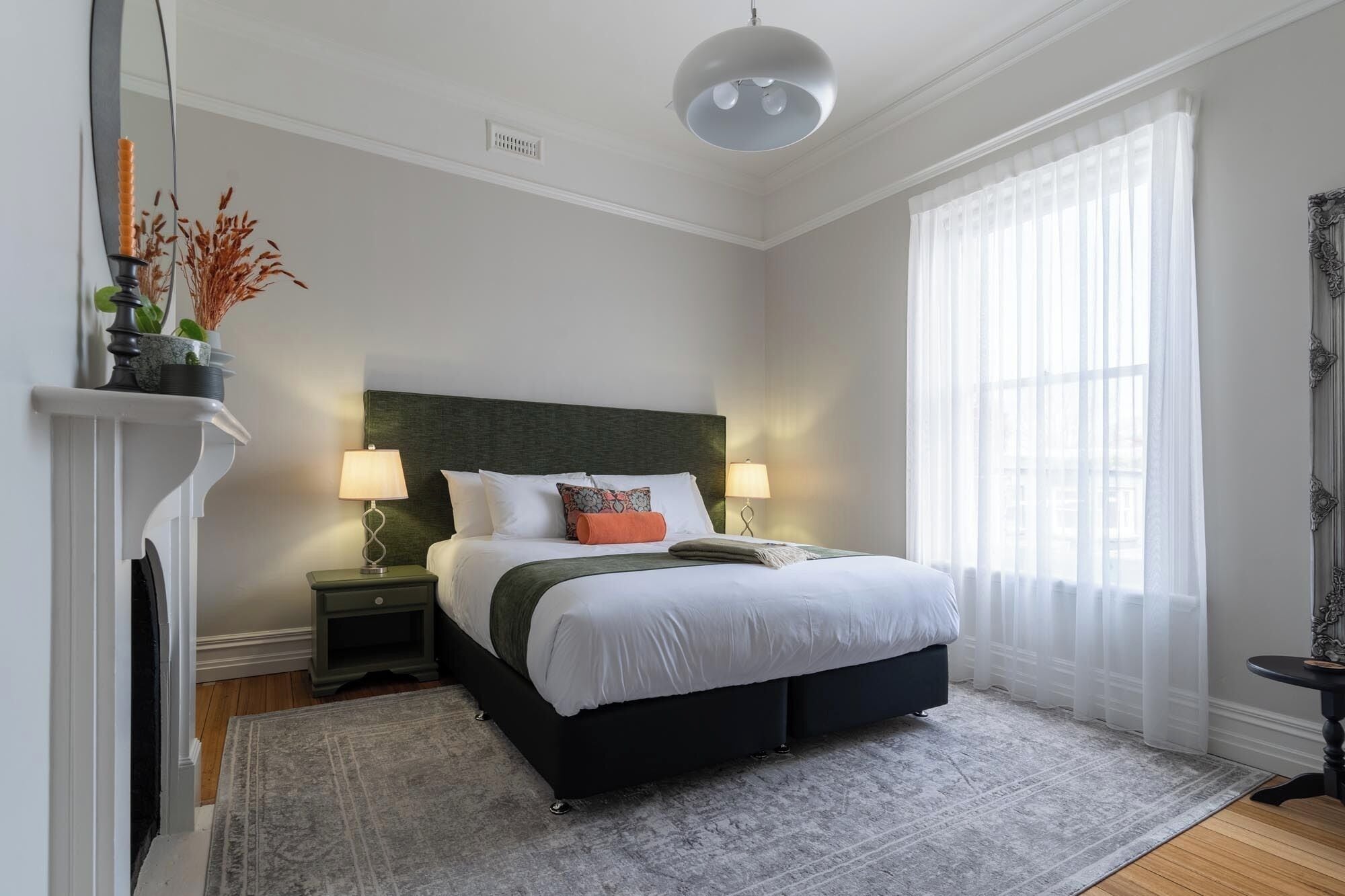 A bedroom with a bed, bedside lamps, a fireplace, and a large window with sheer curtains.