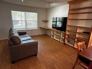 Ein Wohnzimmer mit einer Couch, einem Fernseher auf einem Schrank, einem Holzregal und einem Fenster mit Jalousien.
