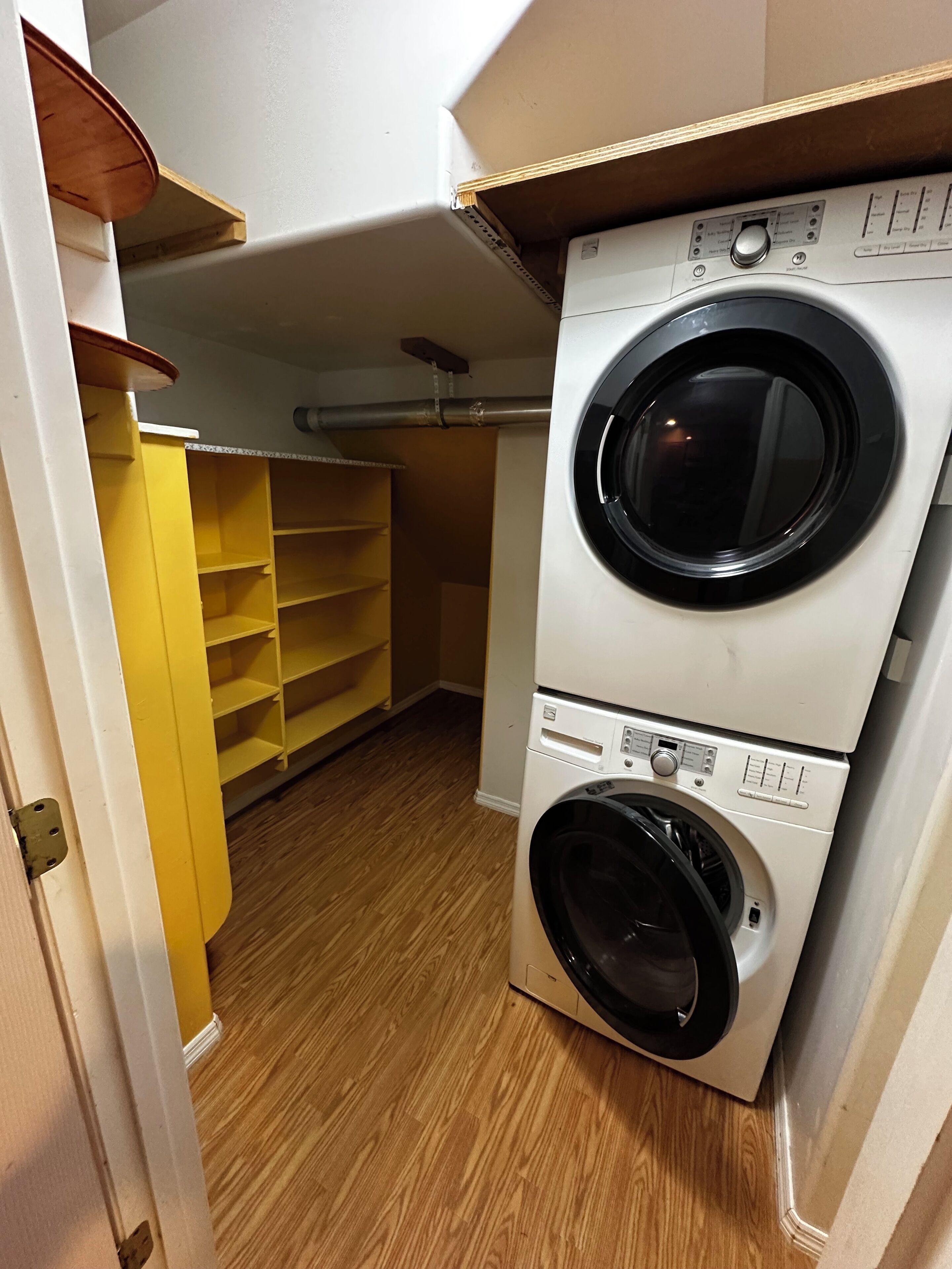 A laundry room with a stacked washer and dryer, wooden flooring, and a yellow shelving unit.