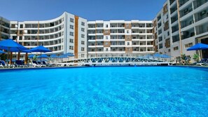 Een hotel met een groot zwembad, meerdere balkons en blauwe parasols.